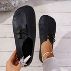 Black flat shoes with decorative buttons held by a hand on a light wooden floor.