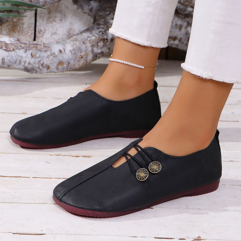 Black loafers with gold buttons worn by a person on a light wooden floor.